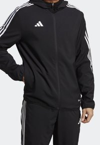 Černá bunda s kapucí a zipem na přední straně, se třemi bílými pruhy na rukávech. Na prsou je logo Adidas.