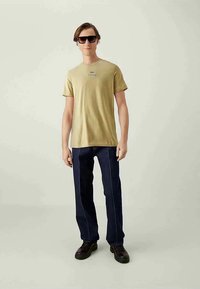 Νεαρός άνδρας που στέκεται, φοράει μπεζ μπλούζα με κοντά μανίκια και λογότυπο, σκούρα μπλε παντελόνι με κάθετες ραφές, μαύρα παπούτσια και σκούρα γυαλιά ηλίου.