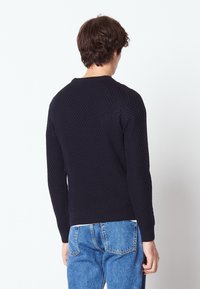 Pull en maille bleu marine avec un design texturé, des poignets et un ourlet côtelés, porté avec un jean bleu clair, vu de dos.