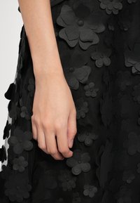 Main reposant à côté d'une jupe noire décorée d'appliques florales noires en couches de différentes tailles sur un tissu transparent.
