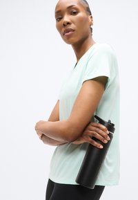 Αθλητική ανοιχτοπράσινη μπλούζα με κοντά μανίκια, σε συνδυασμό με μαύρο παγούρι νερού με καπάκι που ανοίγει προς τα πάνω, που κρατιέται στο μπράτσο.