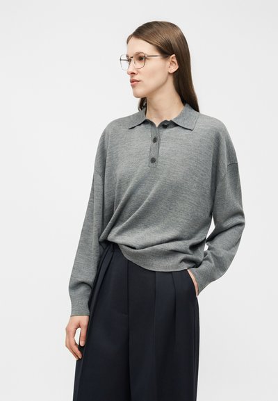 Kobieta o długich brązowych włosach, nosząca okrągłe okulary, luźną szarą koszulę polo z długim rękawem oraz czarne spodnie z szerokimi nogawkami, stojąca na białym tle.