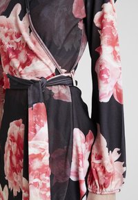 Blommig omlottklänning i lätt tyg, med stora rosa pionmönster på en mörk bakgrund. Inkluderar långa ärmar och ett knyt i midjan.