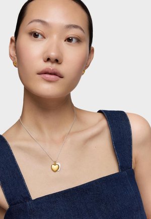 Jeune femme portant des boucles d'oreilles en forme de cœur doré et un collier pendentif assorti, vêtue d'un haut en denim à encolure carrée, regardant légèrement vers la droite sur fond blanc.