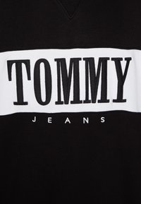 Sudadera negra con una llamativa franja horizontal blanca y letras en relieve negras que dicen "TOMMY" sobre "JEANS" en una fuente blanca más pequeña.