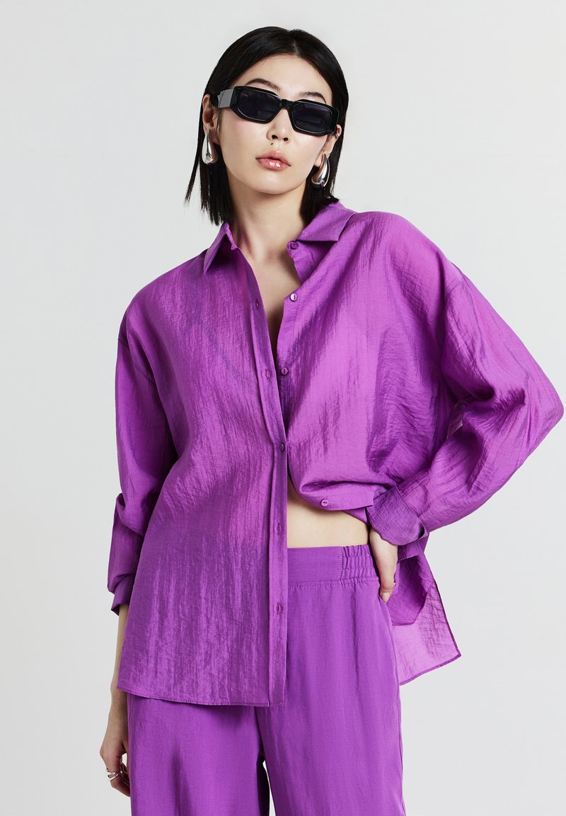 Femme portant une chemise violette surdimensionnée et un pantalon, des lunettes de soleil noires, des boucles d'oreilles en argent, posant avec une main sur la hanche devant un fond blanc.
