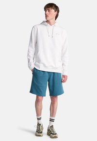 Weißer Baumwoll-Hoodie mit Fronttasche und Kordeln, kombiniert mit türkisfarbenen Shorts. Das Modell trägt beige Sneaker und schwarz-weiße gestreifte Socken.
