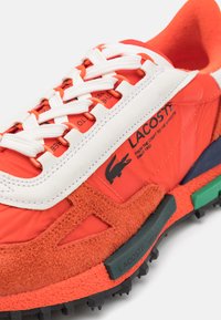 Oransje sneakers med hvite lisser, med en semsket tekstur, gummisåle og kontrasterende grønne og svarte detaljer. Lacoste-logoen er vist.