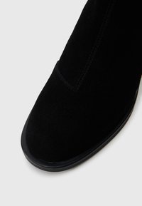 Μαύρη σουέτ μπότες έως τον αστράγαλο με στρογγυλή μύτη, διαθέτει λεία υφή και χαμηλή μαύρη καουτσούκ σόλα. Οι λεπτομέρειες ραφή�ς είναι ορατές.