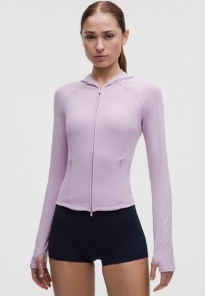 Vrouw draagt een aansluitende lichtpaarse hoodie met rits en duimgaten en zwarte sportshorts tegen een effen achtergrond.
