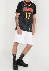 Persoon in een zwart Atlanta 17 basketbalt-shirt, witte shorts, zwarte gemarmerde sokken en zwarte sneakers, staande tegen een witte achtergrond.