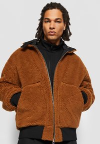 Veste en polaire marron avec fermeture éclair, deux poches avant et doublure intérieure noire. Le tissu a une apparence texturée et douce.