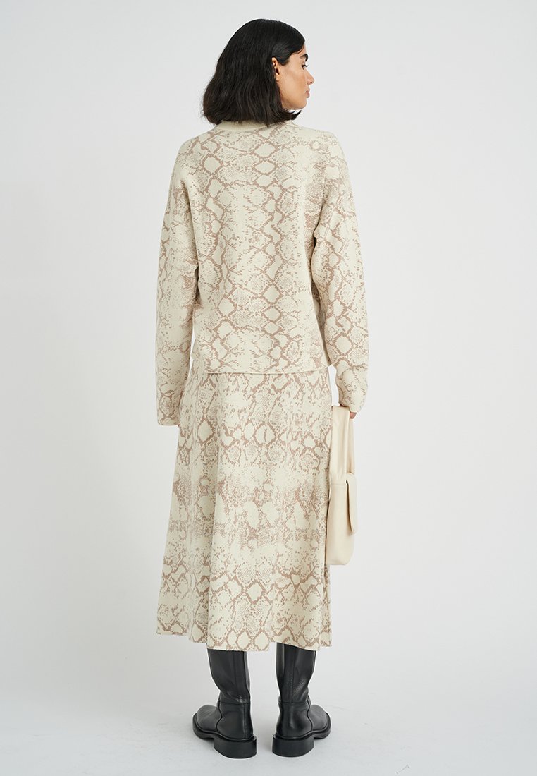 Langes, beiges Kleid mit Schlangenprint-Design, ausgestattet mit langen Ärmeln und einer lockeren Passform. Kombiniert mit einer cremefarbenen Handtasche und schwarzen knielangen Stiefeln.