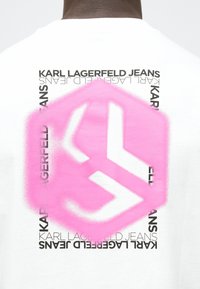 Vit t-shirt med en stor rosa sexhörnig spraymålad logotyp och svart text med orden "KARL LAGERFELD JEANS." Mjuk bomullstyg.