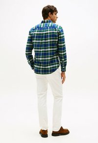 La camisa de cuadros presenta tonos verdes y azules en una tela texturada, con un cuello tipo button-down y mangas largas. Se combina con pantalones blancos y zapatos marrones.