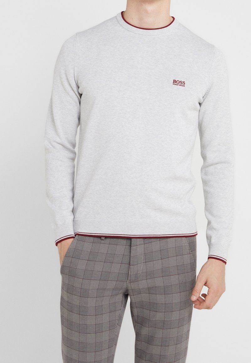 Man in een lichtgrijze sweater met lange mouwen en rode details, voorzien van het Hugo Boss-logo, gecombineerd met grijs geruite broek, met ontspannen handen langs zijn zij.