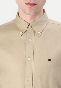 Man in een beige knoopshirt met button-down kraag en een klein rechthoekig rood, wit en blauw logo op de linkerborst.