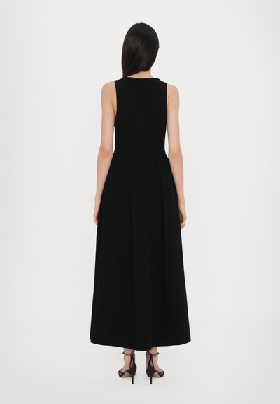 Zwarte, mouwloze maxidress met een getailleerd lijfje en een uitlopende rok, voorzien van een gladde textuur en minimale details. G gedragen met zwarte hakken.