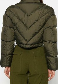 Zelená prošívaná bunda s krátkým střihem a vzorem ve tvaru šipky. Má elastický lem a plyšovou texturu. Nositelná s odpovídajícími zelenými kalhotami.