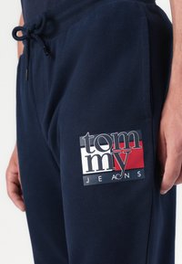 Marineblauwe sweatpants van zachte stof met een trekkoord in de taille, voorzien van een groot logo-embleem in rood, wit en zwart aan de zijkant.