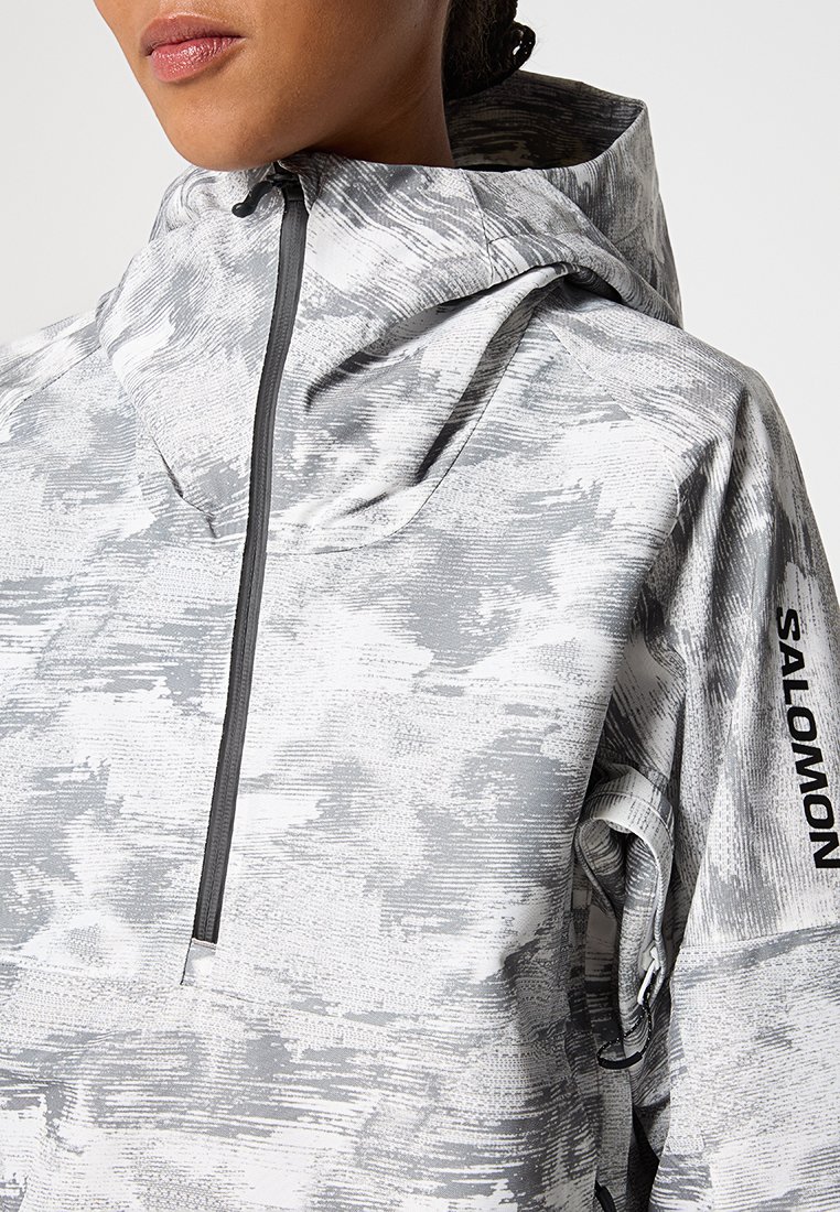 Persona con una chaqueta con capucha de patrón gris y blanco, con cremallera frontal y "SALOMON" impreso en la manga.