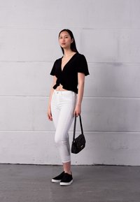 Top preto com nó de mangas curtas, jeans brancos até os tornozelos, tênis pretos e uma pequena bolsa texturizada em frente a uma parede cinza.