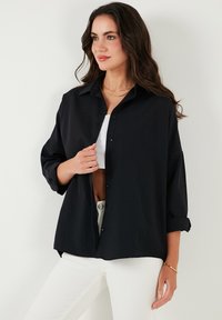 Camicia nera con bottoni, maniche lunghe, vestibilità rilassata e polsini arrotolati, abbinata a un top corto bianco e pantaloni di colore chiaro.