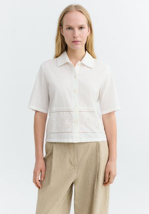 Frau trägt ein weißes, kurzärmeliges Hemd mit Knöpfen, verziert mit Stickereien von Fisch-Ausschnitten, und beige Hosen mit weitem Bein, stehend vor einem einfarbigen Hintergrund.