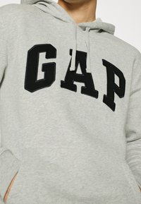 Felpa grigia con tasca a canguro, con un grande logo "GAP" in lettere spesse e ricamate di colore nero. Include il cappuccio regolabile con cordini.