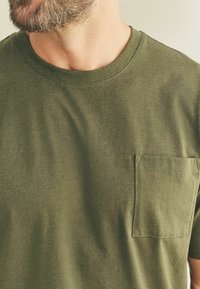 Olivgrünes Baumwoll-T-Shirt mit Rundhalsausschnitt und einer Brusttasche. Weiche Textur mit dezenten Nähdetails am Ausschnitt und der Tasche.