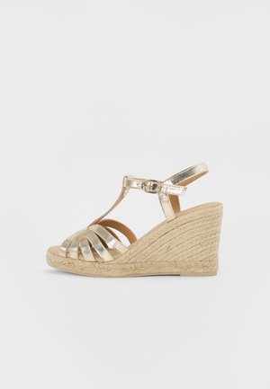 Wedge sandaal met een metallic gouden bovenkant, gekruiste banden, een T-strap en een geweven jute tussenzool voor textuur en hoogte.