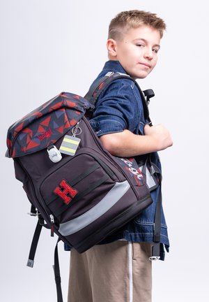 Junge trägt eine dunkle Jeansjacke und beige Hose und trägt einen großen schwarzen und roten Rucksack mit geometrischem Muster auf einer Schulter.