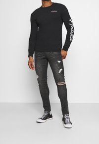 Svart långärmad skjorta med vitt logga på bröstet och ärmen, matchad med mörka slitna skinny jeans och svarta hög-top sneakers.