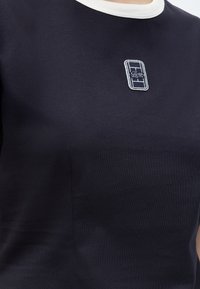 Κοντινή λήψη σκούρου μπλε πουκαμίσου με λευκό γιακά και μικρό ορθογώνιο έμβλημα με πλέγμα και λογότυπο κροκοδείλου στο στήθος.