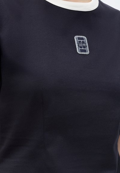 Gros plan d'une chemise bleu marine avec un col blanc et un petit patch rectangulaire comportant une grille et un logo crocodile sur la poitrine.