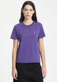 T-shirt di cotone viola con scollatura rotonda e maniche corte, caratterizzata da un piccolo logo bianco sul lato sinistro del petto. Vengono indossati pantaloni neri.