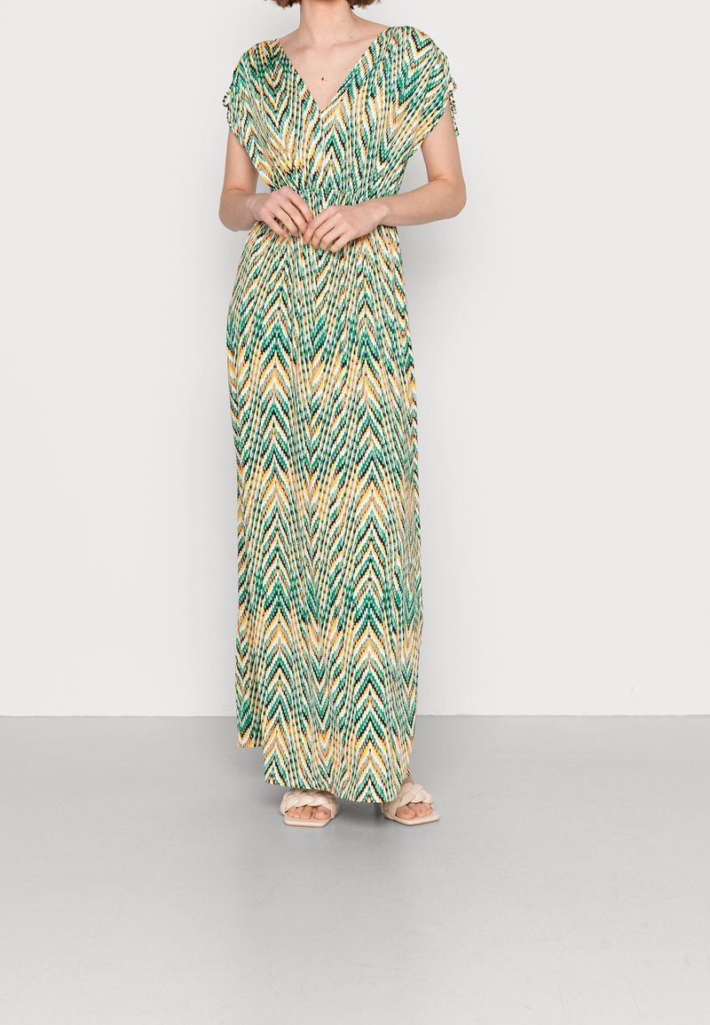 Femme portant une longue robe à manches courtes avec un motif zigzag multicolore et une taille froncée, debout sur un sol gris devant un mur blanc.