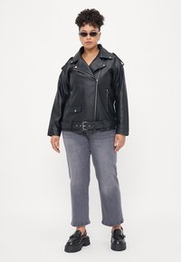 Black faux leather biker jacket with zippered pockets, silver hardware, and a belted waist. Worn over gray straight-leg jeans and chunky black shoes.
