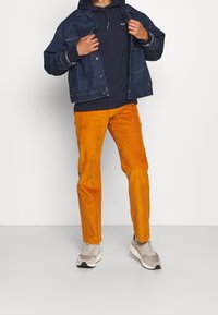 Giacca di jeans, felpa con cappuccio blu scuro e pantaloni in velluto a coste arancioni. La giacca presenta bottoni e una vestibilità comoda. I pantaloni hanno una trama a costine verticali. Scarpe da ginnastica neutre.