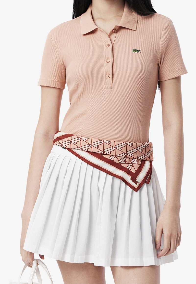 Femme portant un polo rose clair avec un logo vert, une jupe plissée blanche et une écharpe à motifs nouée autour de la taille.