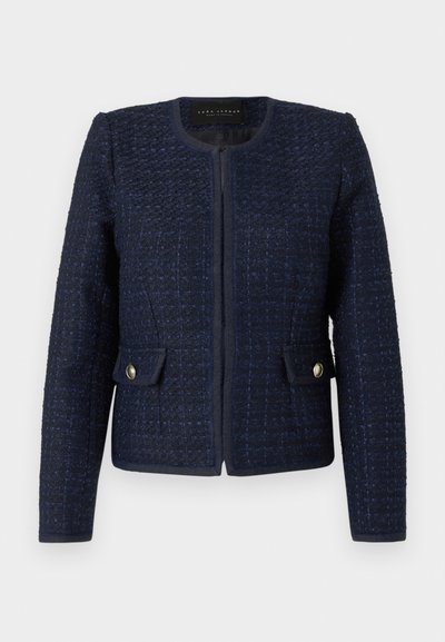 Giacca di tweed blu navy con una forma aderente, collo rotondo e chiusura frontale con zip. Presenta due tasche con bottoni dorati. Tessuto texturizzato.