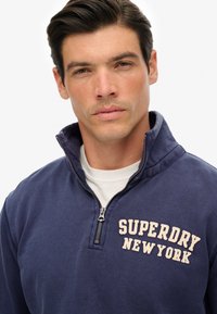 Granatowa, zapinana na zamek bluza z bawełny o miękkiej strukturze, z dużym kremowym logo "SUPERDRY NEW YORK" na piersi.