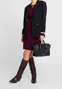 Musta nahkainen käsilaukku kultaisilla yksityiskohdilla, yhdistetty burgundinpunaisen mekon, mustan bleiserin ja ruskeiden polvenkorkuisten tekstiilisaappaiden kanssa.