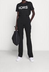 Musta T-paita, jossa valkoinen "KORS"-logo, yhdistetty mustiin housuihin. Malli pitää mustaa olkalaukkua ja käyttää valkoisia tennareita. Minimalistinen design.