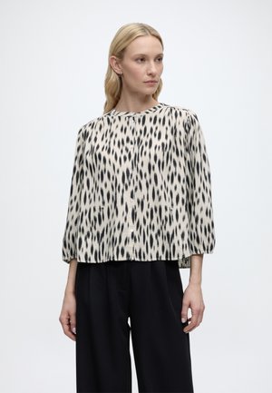 Kobieta ubrana w beżową bluzkę w czarne pionowe kropki, z rękawami o długości do łokcia, oraz czarne szerokie spodnie, stojąca na jednolitym tle.