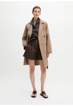 Frau trägt beigefarbenen Trenchcoat über dunkelbraunem Hemd mit Knöpfen und Shorts, dazu schwarze Sandalen, steht vor schlichter weißer Wand.