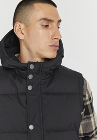 Čierny puff vest s vysokým golierom, so striebornými patentkami a mäkkou textúrou. Nosí sa cez károvanú flanelovú košeľu.