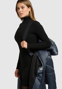 Mustast soonikust kootud kleit kõrge kraega, kombineeritud sinise paksu jopega. Jopel on läikiv kangas ja rihmadel logomustrid.