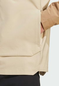 Beige lichtgewicht jack met een matte afwerking, voorzien van een voorzak en een losse pasvorm. De mouw heeft een contrasterende taupekleurige manchet.