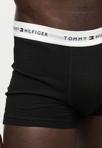 Primo piano di una persona che indossa boxer neri Tommy Hilfiger con una fascia in vita bianca con il logo e la bandiera del marchio.